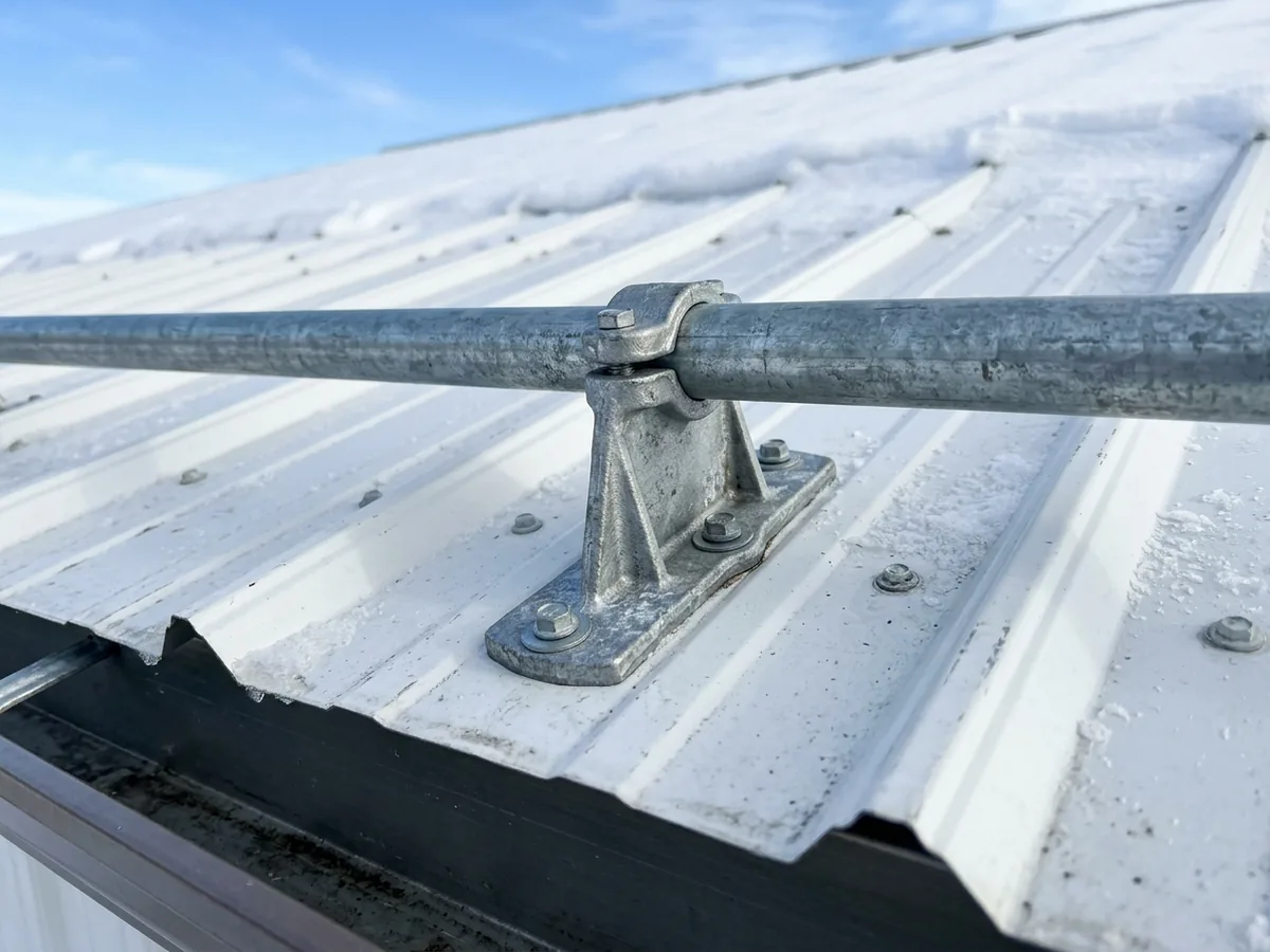 Steel snow guard bracket screwed through a ribbed metal roof panel near the eave, holding a round pipe